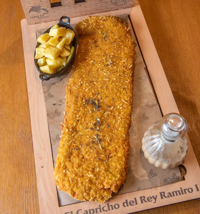 Cachopo sobre bandeja de madera junto a una cazuelina con patatas fritas y un tarro con salsa